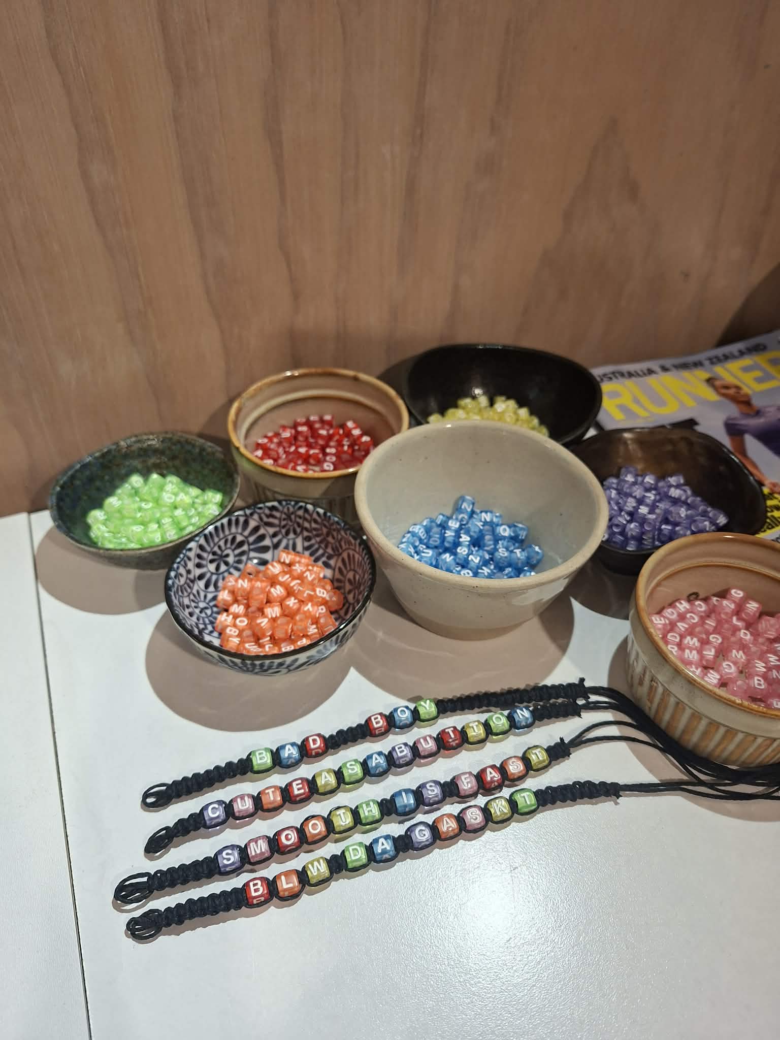 Handmade word bracelets made from colourful letter beads on cord, with bowls of red, green, orange, blue, purple, yellow and pink beads behind them